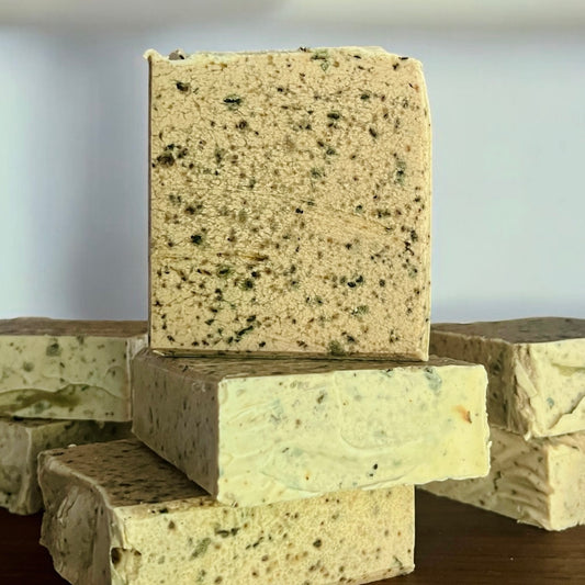 Une pile de barres rectangulaires de savon exfoliant MAGDA thé vert & lait de coco de La Sultana Savonnerie dégage un charme naturel avec son infusion d'herbes mouchetées de vert et de brun. Les barres beige clair présentent une texture rugueuse sur les côtés et une surface plus lisse, le tout sur fond de mur blanc immaculé.