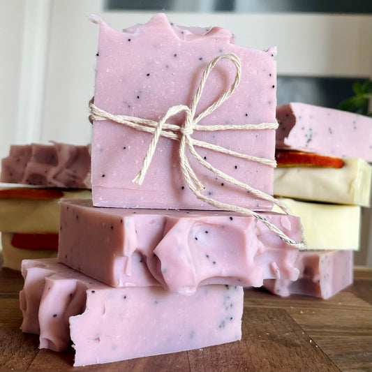 Plusieurs morceaux de savon artisanal sont empilés sur une surface en bois. Les pains de savon sont roses avec des taches et des bords rugueux. Le top bar, un Savon exfoliant LILI graines de pavot, jojoba et pamplemousse de La Sultana Savonnerie | Savons et soins naturels faits main | Québec, est lié par une fine ficelle. D’autres pains de savon de différentes couleurs et couches se trouvent en arrière-plan.