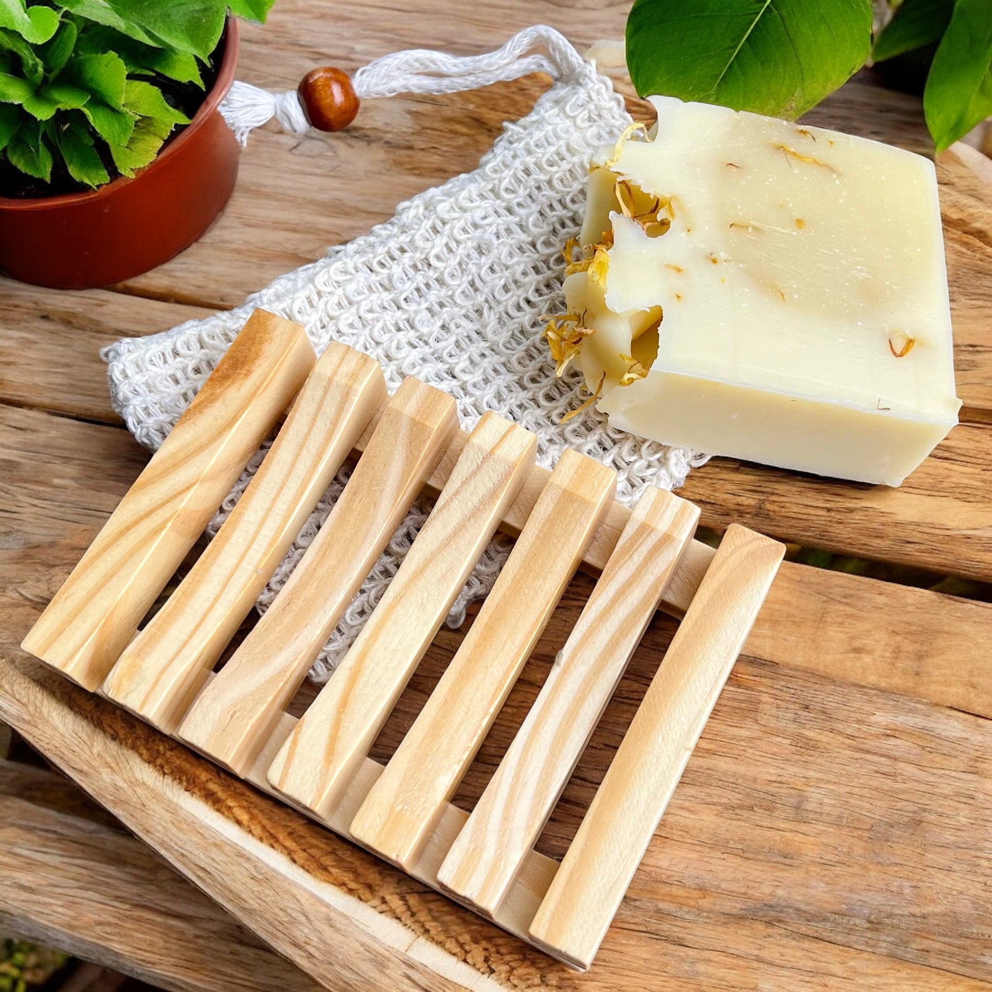 A Porte savon et pochette en sisal de La Sultana Savonnerie | Savons et soins naturels faits main | Le Québec, fabriqué à partir de bois de pin, est posé sur une surface en bois à côté d'un morceau de savons artisanaux aux pétales de fleurs séchées. Un sac de gommage tricoté et une petite plante en pot sont également visibles en arrière-plan.