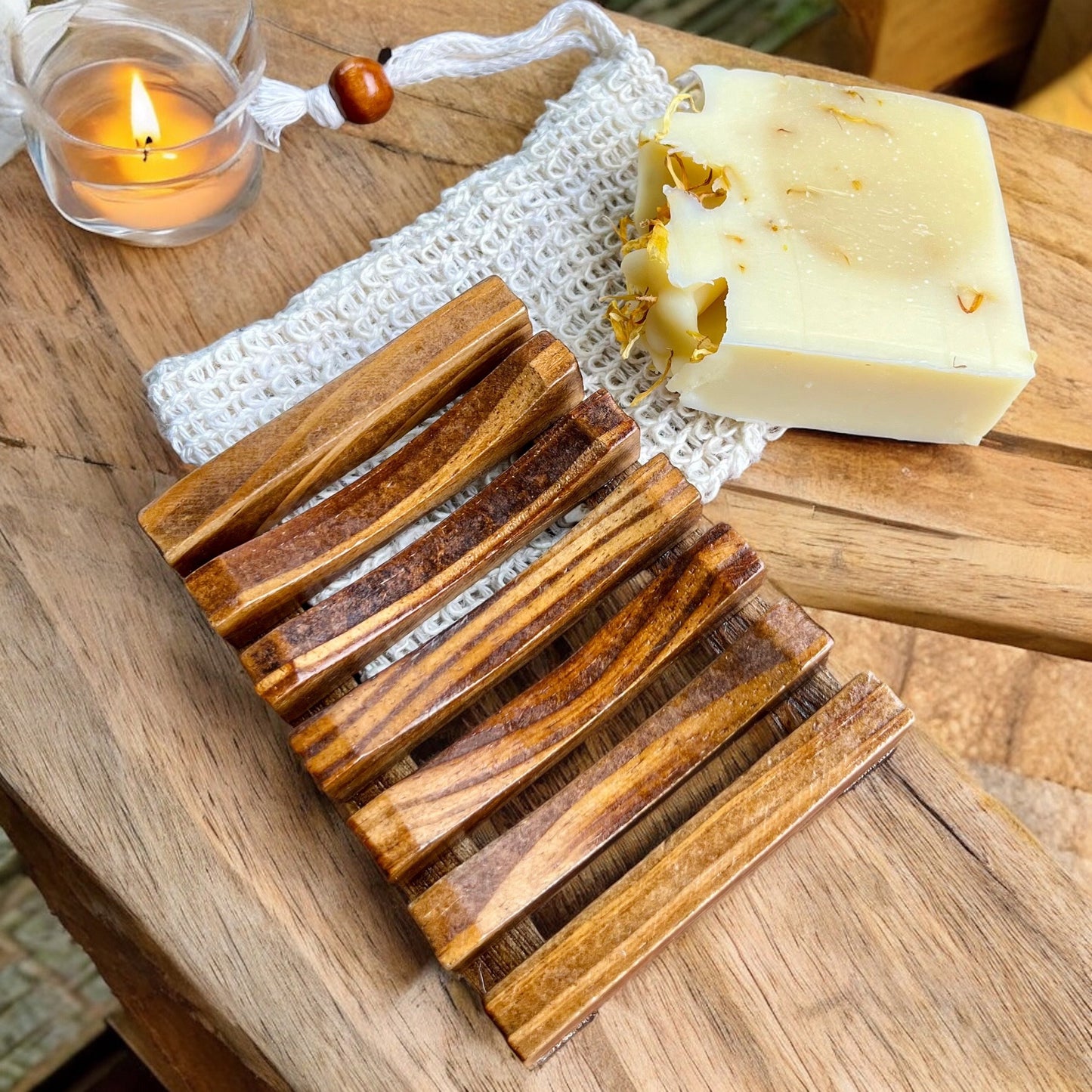 Un **Porte-savon et pochette en sisal** de **La Sultana Savonnerie | Savons et soins naturels faits main | Québec** en bois de pin latté est posé sur une surface en bois à côté d'une barre de savons artisanaux partiellement utilisés avec des pétales de fleurs. Derrière eux, une bougie allumée est placée dans un petit support en verre et une pochette à cordon blanc est partiellement visible.