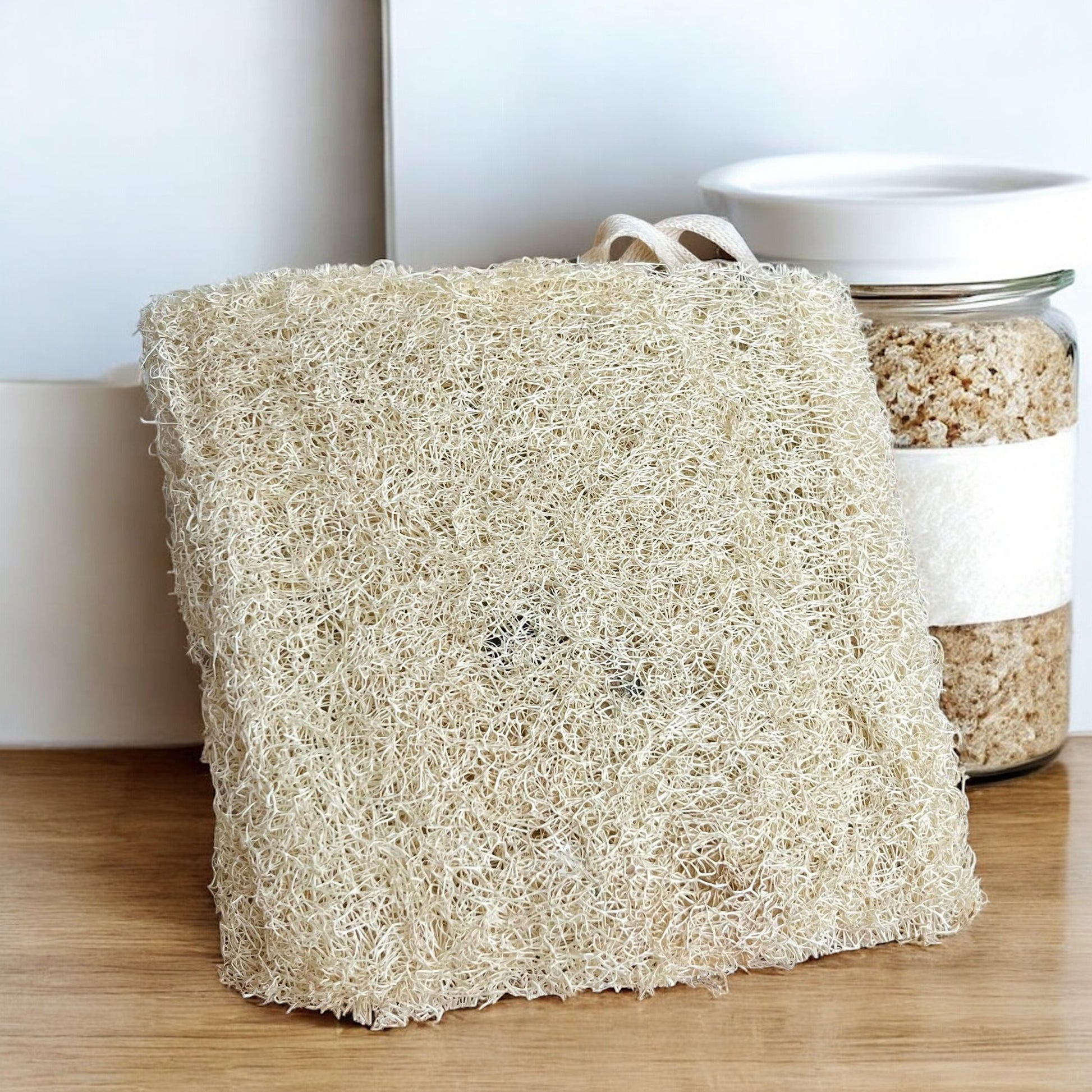 Une Grande éponge Loofah naturelle beige et fibreuse de La Sultana Savonnerie | Savons et soins naturels faits main | Québec, à laquelle est attachée une ficelle blanche, est placée sur une surface en bois à côté d'un pot en verre rempli de sels de bain granuleux. Le décor présente un mur blanc et des surfaces angulaires supplémentaires en arrière-plan, mettant en valeur cet outil d'exfoliation douce idéal.