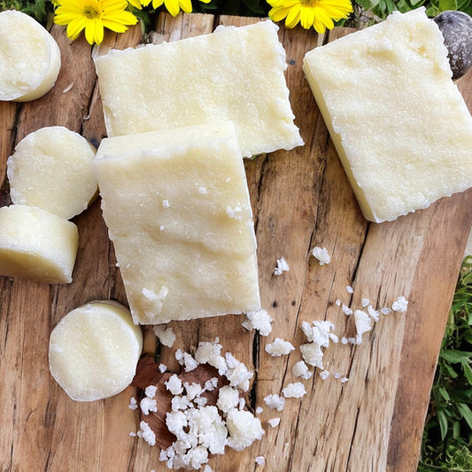 Une surface en bois présente quatre barres rectangulaires de l'Exfoliant MUSSASA au sucre visage et corps de La Sultana Savonnerie | Savons et soins naturels faits main | Québec, trois petits morceaux de savon ronds et quelques morceaux de savon émiettés. Plusieurs fleurs jaunes sont disposées près des barres de savon.