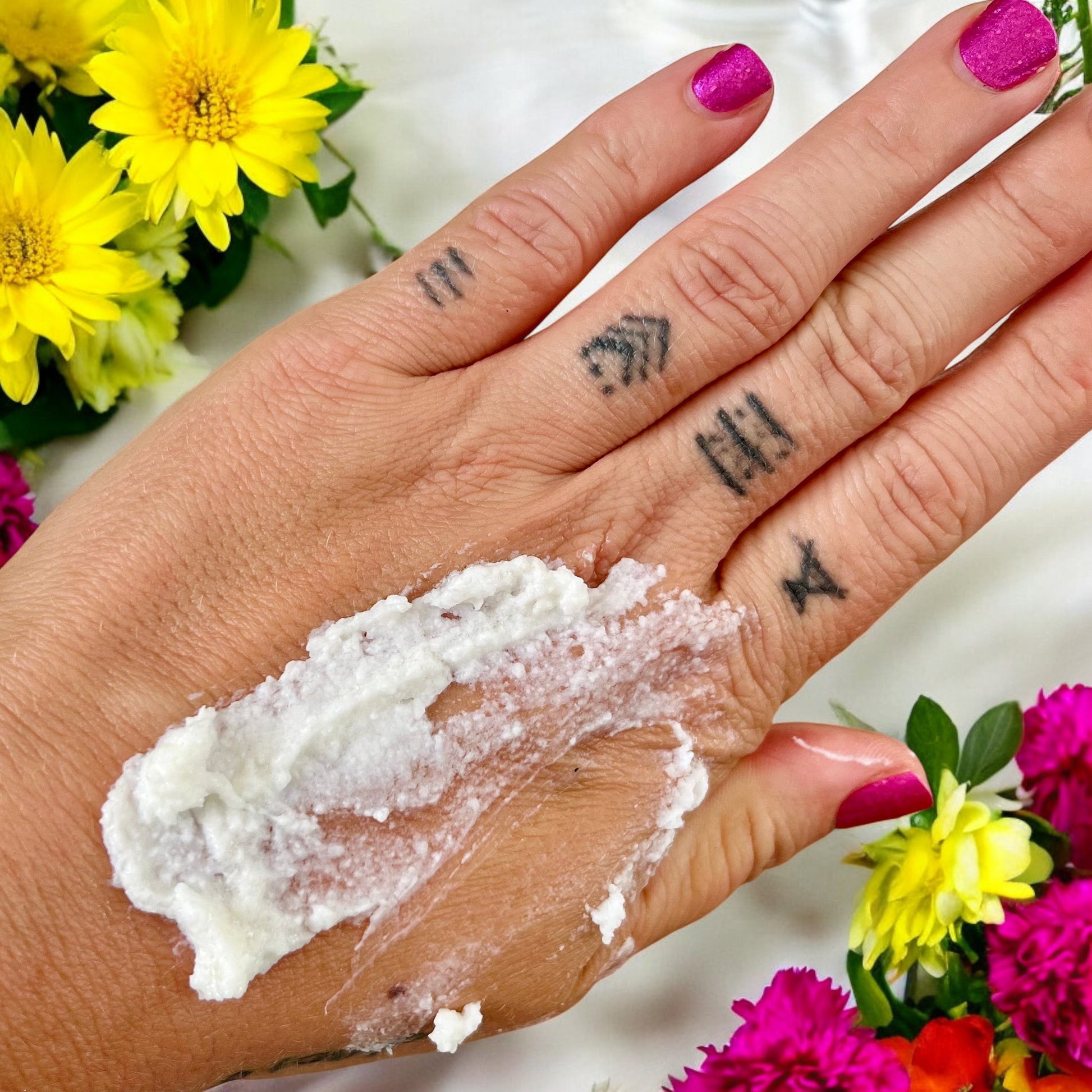 Gros plan d'une main avec la Crème hydratante ÉLOÏSE hydrolat de lavande appliquée sur le dos, mettant en valeur plusieurs tatouages sur les doigts. Les ongles sont peints en rose vif, et il y a des fleurs jaunes et roses en arrière-plan, mettant l'accent sur la réparation et l'apaisement. Ce produit luxueux vous est proposé par La Sultana Savonnerie | Savons et soins naturels faits main | Québec.