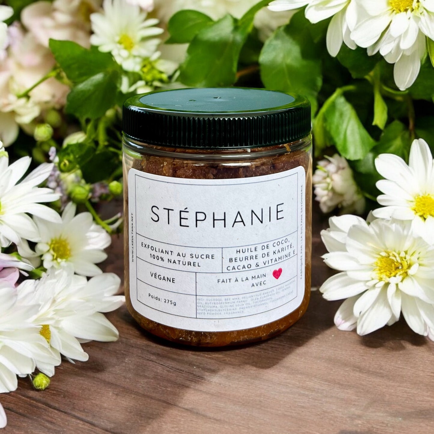 Un pot d'Exfoliant corporel STÉPHANIE chocolat et vanille de La Sultana Savonnerie, un gommage au sucre exfoliant 100% naturel, est posé sur une surface en bois entourée de fleurs blanches. L'étiquette indique qu'il est végétalien, fait à la main et contient de l'huile de coco, du beurre de karité, de la vitamine E et du cacao pour régénérer la peau. Le pot pèse 275 g.