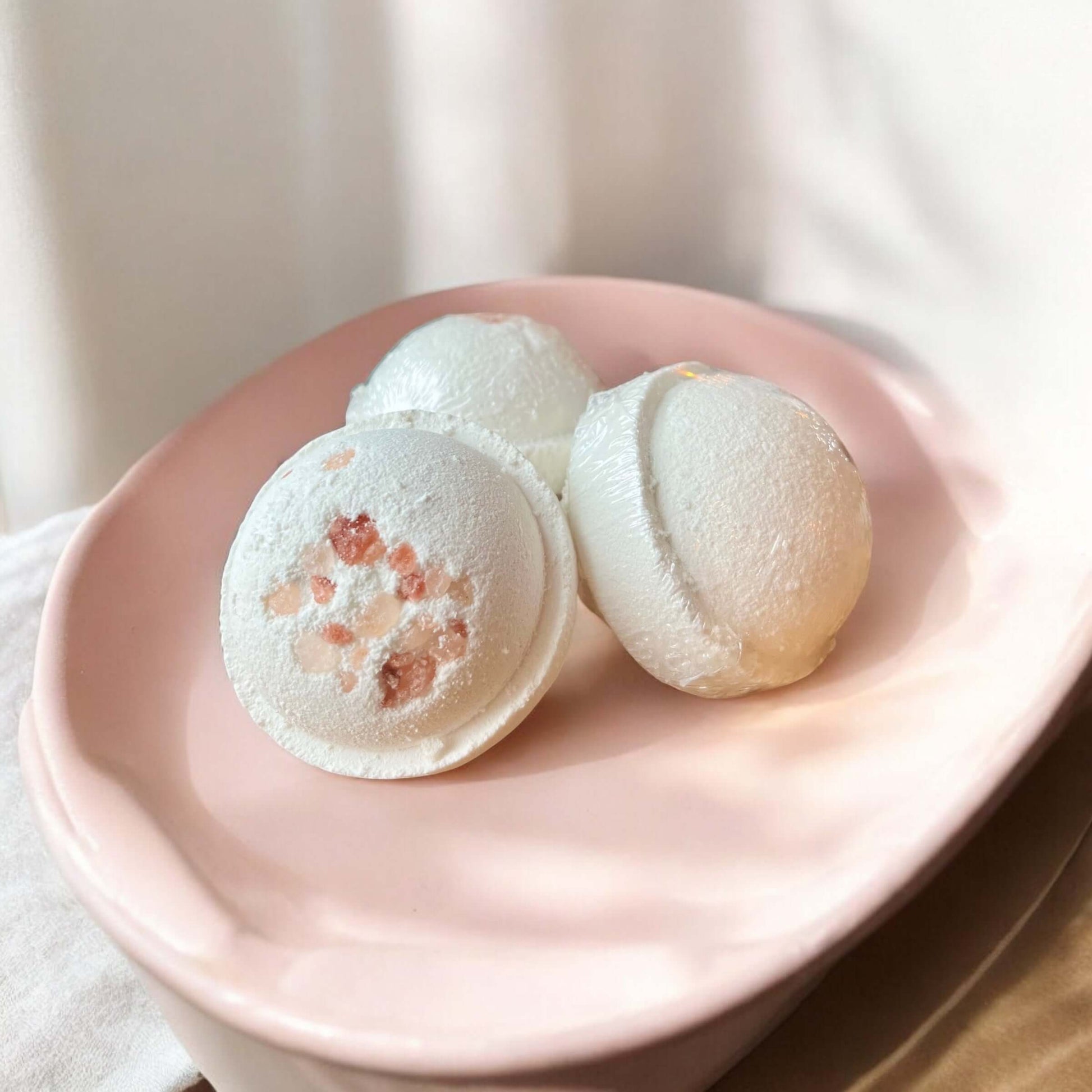 Trois bombes de bain rondes et blanches, connues sous le nom de Bombe de bain ALEXANDRE à la menthe poivrée de La Sultana Savonnerie, sont posées sur un plat en céramique rose. L'une des bombes de bain, infusée d'ingrédients naturels, présente des cristaux de sel roses incrustés à sa surface. Le plat est placé sur une surface de couleur claire avec un tissu blanc doux en arrière-plan. La Sultana Savonnerie se spécialise dans les savons naturels faits à la main et les produits de soins de la peau du Québec.