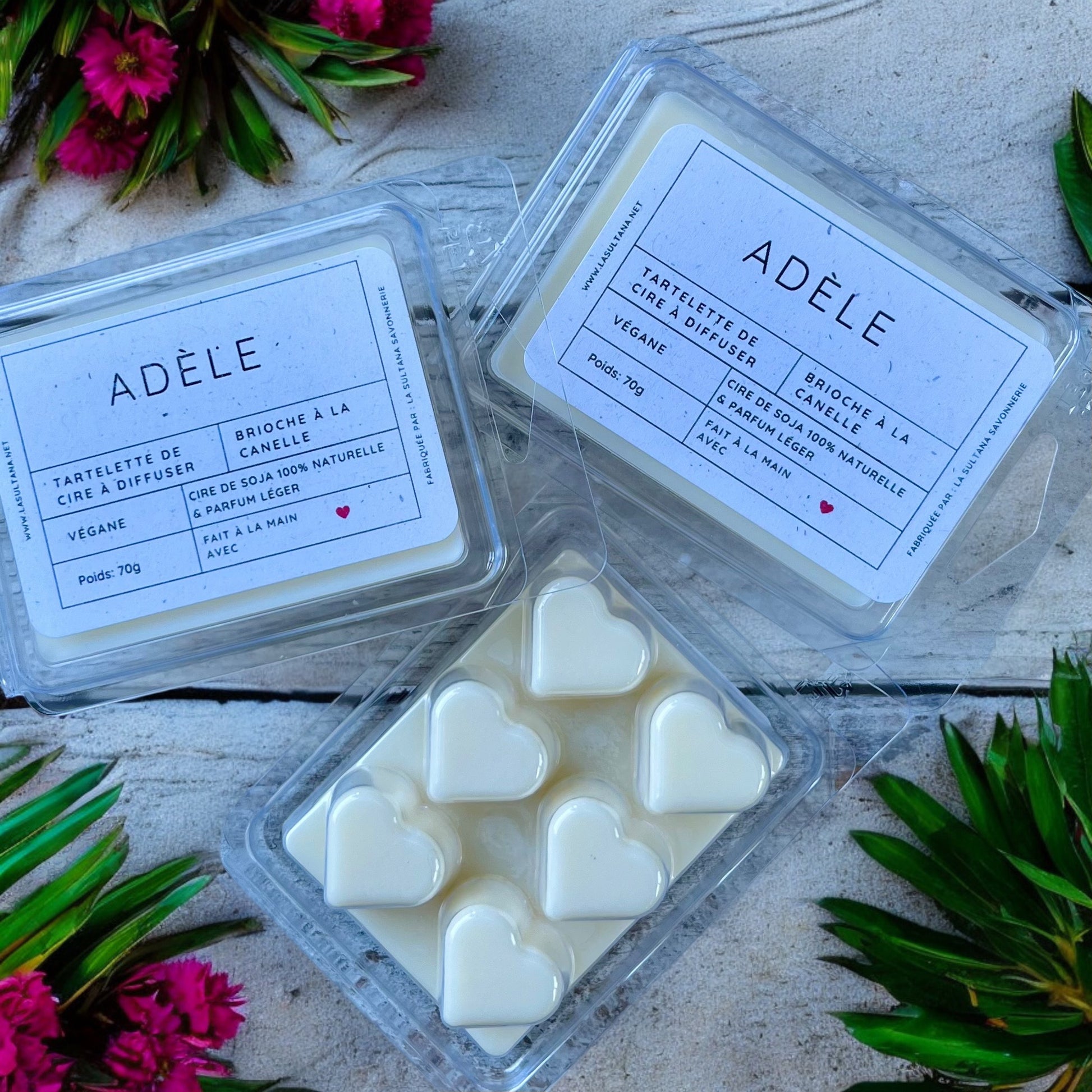 Une photographie présente trois paquets de fondants de cire « ADÈLE » de La Sultana Savonnerie. Un paquet ouvert révèle six fondants blancs en forme de cœur. Le texte de l'emballage indique qu'ils sont végétaliens et naturellement parfumés à la brioche à la cannelle. Des fleurs roses décorent l'arrière-plan.