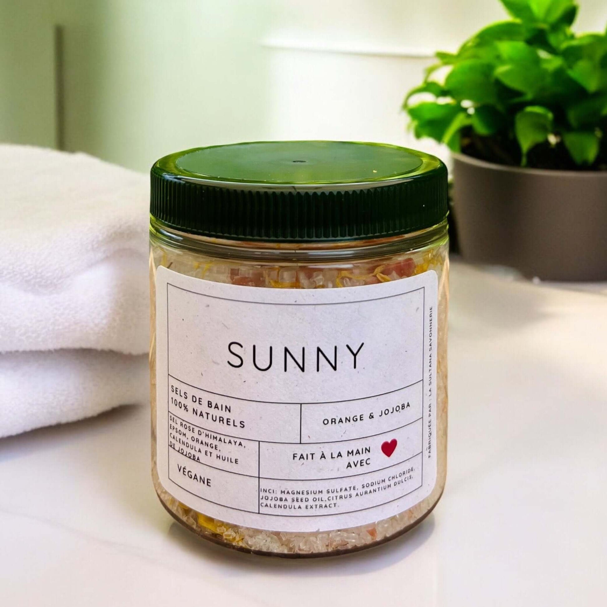 Un pot étiqueté « Sels de bain SUNNY orange sucrée et calendula » est posé sur une surface blanche. Le pot a un couvercle vert et l'étiquette mentionne « Handmade with Love » par La Sultana Savonnerie | Savons et soins naturels faits main | Québec, et « Vegan ». Une serviette blanche roulée et une plante verte en pot sont à l'arrière-plan.