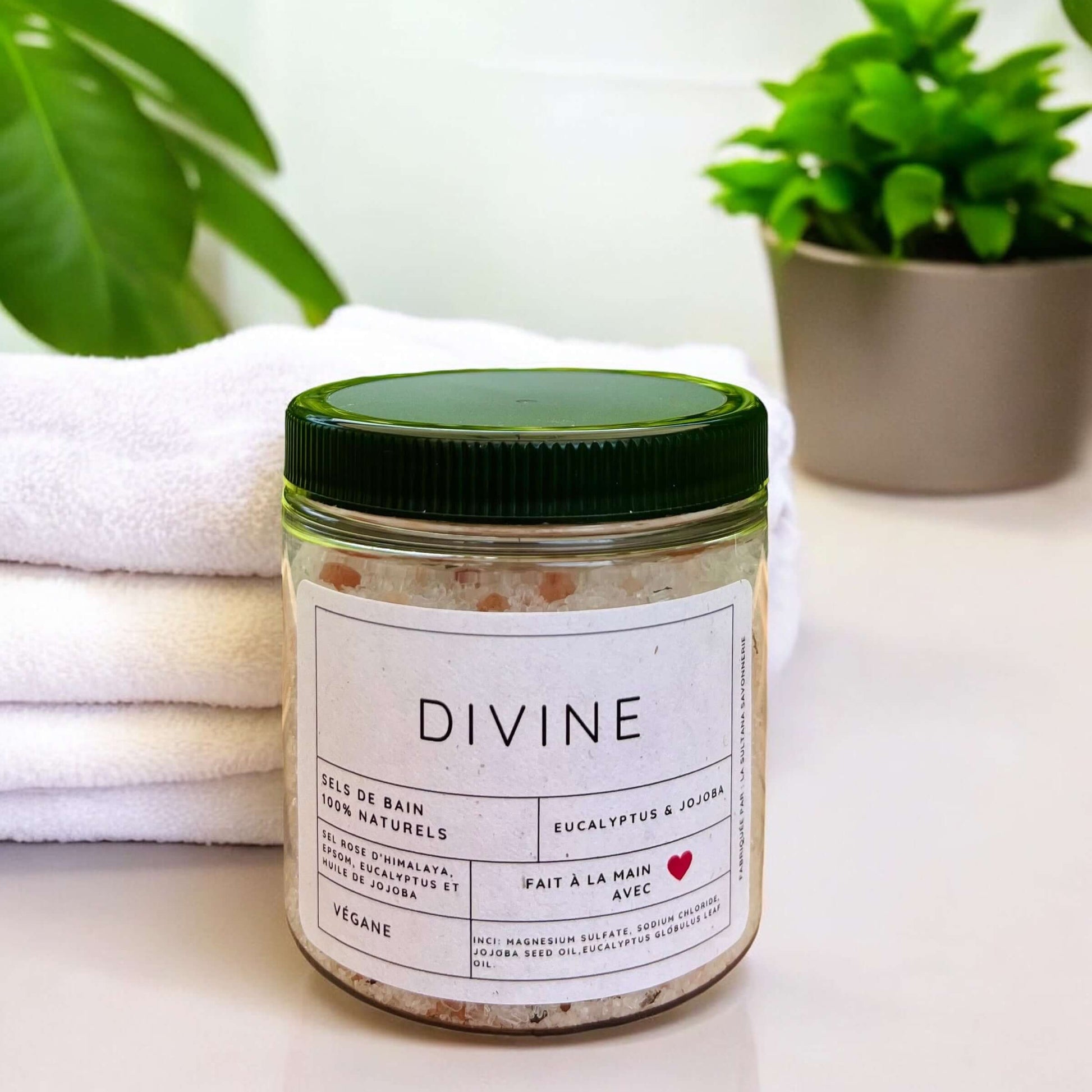 Un pot étiqueté « Sels de bain DIVINE eucalyptus et menthe » de La Sultana Savonnerie, connue pour ses savons naturels et ses produits de soins de la peau faits à la main au Québec, est placé sur une surface blanche. Derrière le pot se trouvent des serviettes blanches soigneusement pliées et une plante verte en pot. Une autre plante à l'arrière-plan est floue. Le pot a un couvercle vert et présente une étiquette au design minimaliste.