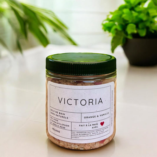 Un pot étiqueté « Sels de bain VICTORIA argile rose, orange et vanille » de La Sultana Savonnerie contient des sels de bain naturels au parfum d'orange et de vanille. L'étiquette souligne sa qualité artisanale, son caractère sans gluten et végétalien, ainsi que l'inclusion de sel d'Epsom. Le pot est posé sur une surface avec une plante verte en arrière-plan.