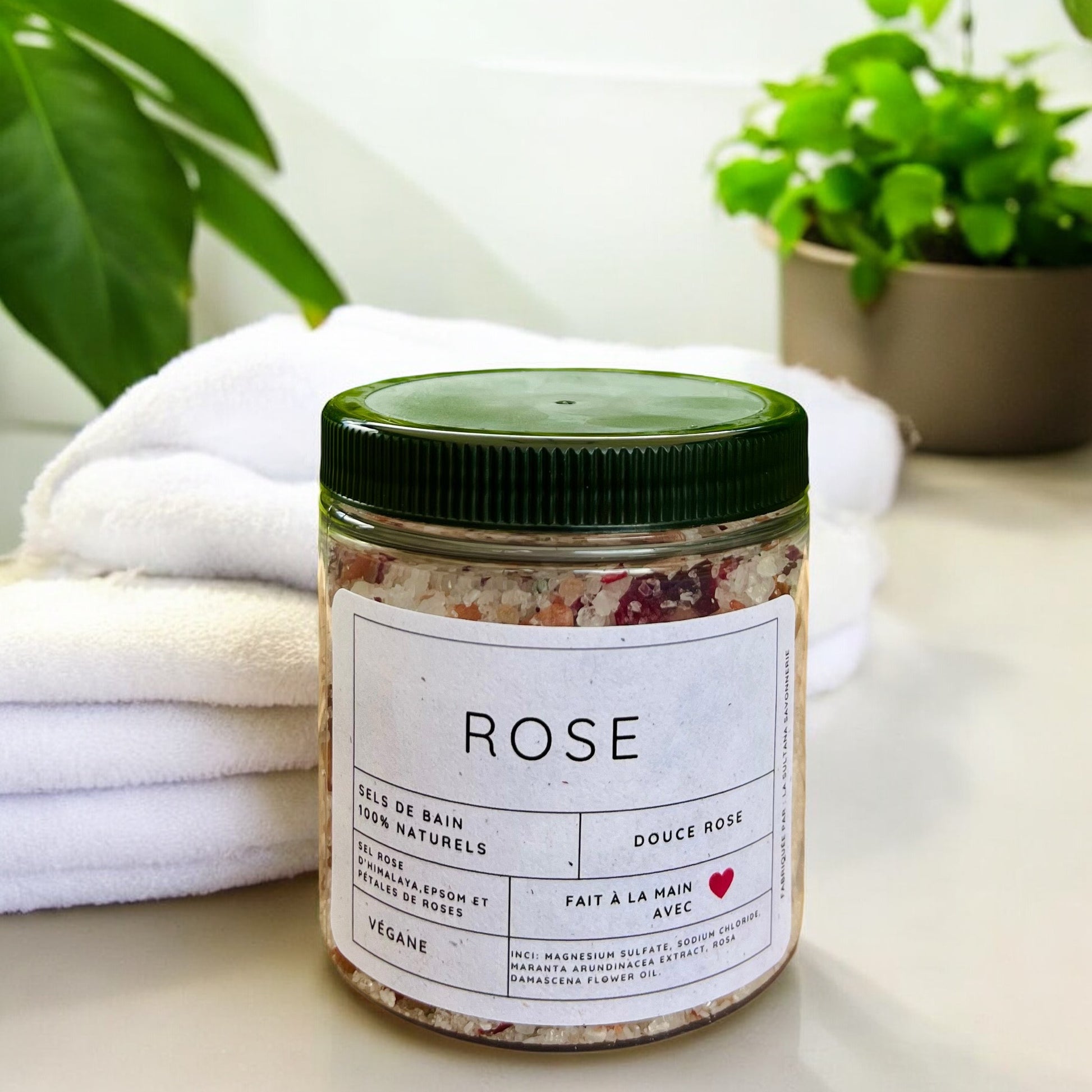 Un pot étiqueté « Sels de bain ROSE rose apaisante » de la marque La Sultana Savonnerie | Savons et soins naturels faits main | Québec est posé sur une surface avec des serviettes blanches empilées et des plantes vertes en pot en arrière-plan. L'étiquette indique que le produit est fait à la main, végétalien et fabriqué avec des ingrédients naturels comme le sel rose d'Himalaya. Le pot a un couvercle vert.