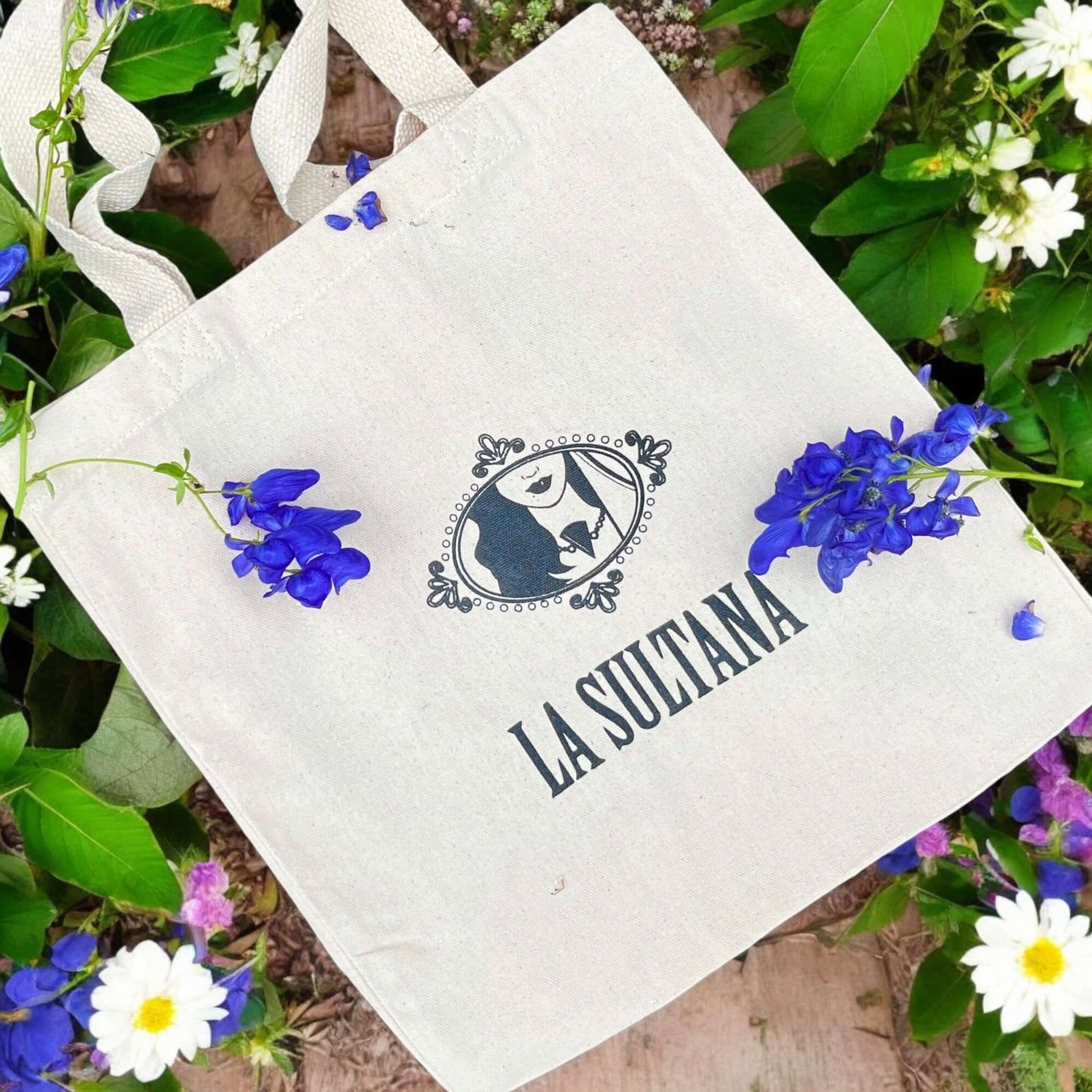 Le « Sac fourre-tout en coton recyclé » de La Sultana Savonnerie | Savons et soins naturels faits main | Québec présente un logo illustré d'une femme élégante et le texte « La Sultana ». Ce sac fourre-tout en toile, posé au milieu de fleurs bleues, violettes et blanches éclatantes, est fabriqué à partir de coton recyclé avec deux poignées. Le fond est composé de feuilles vertes et de planches de bois.