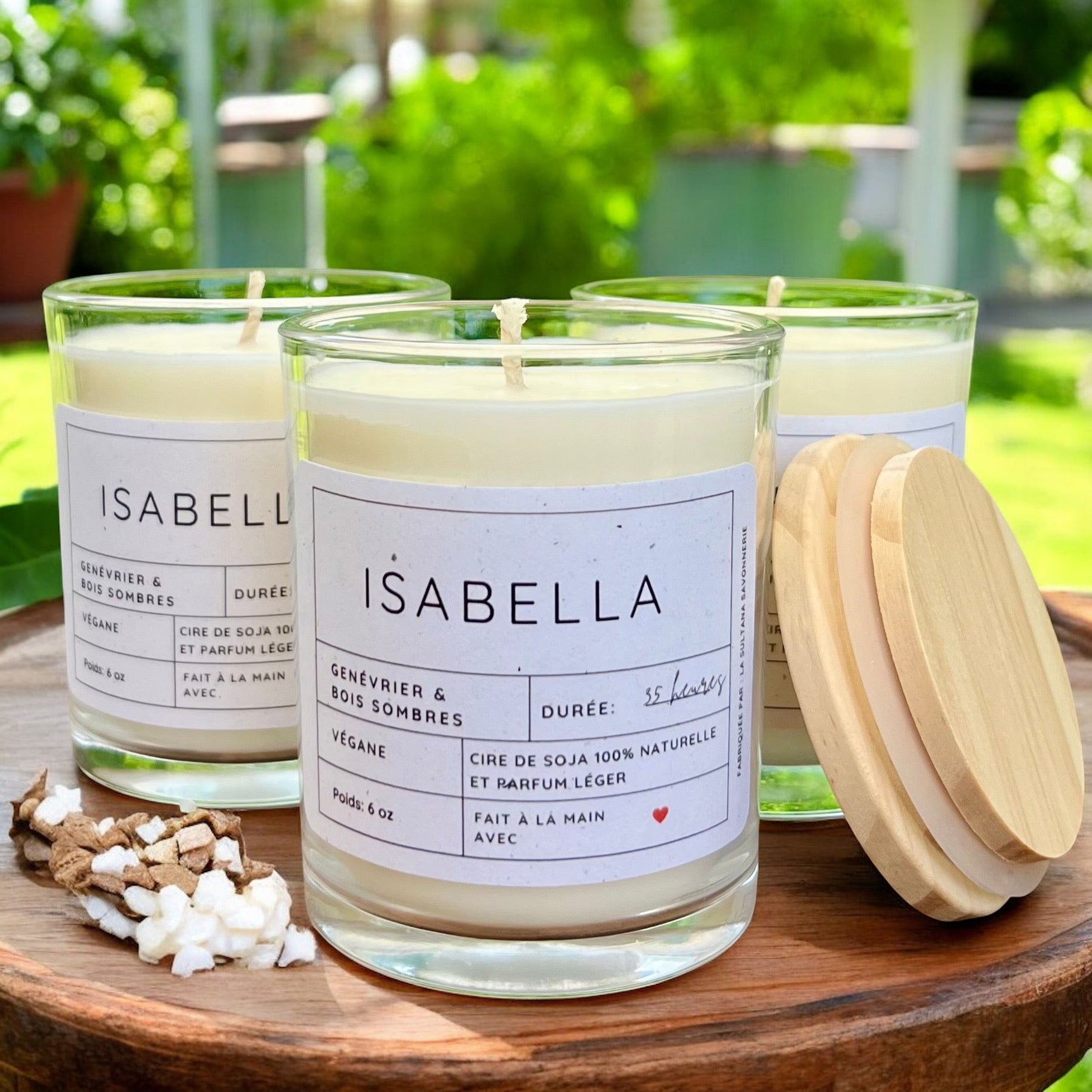 Trois bougies blanches étiquetées dans des pots en verre placés sur une table en bois à l'extérieur. Les étiquettes indiquent « Bougie de soja ISABELLA genévrier et bois sombres » et précisent « Végétalien », « Cire de soja 100 % naturelle » et un parfum « Genévrier et bois sombres » d'une durée de 55 heures. Des couvercles en bois et de petits copeaux de bois sont à proximité, fabriqués par La Sultana Savonnerie | Savons et soins naturels faits main | Québec.