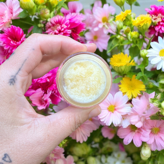 Une main tatouée tient un petit pot de Masque exfoliant pour les lèvres SAKURA miel & citron de La Sultana Savonnerie | Savons et soins naturels faits main | Québec, contenant une substance granuleuse jaune pâle à base d'ingrédients naturels. En arrière-plan, on peut voir un assortiment de fleurs colorées, notamment des fleurs roses, jaunes et blanches.