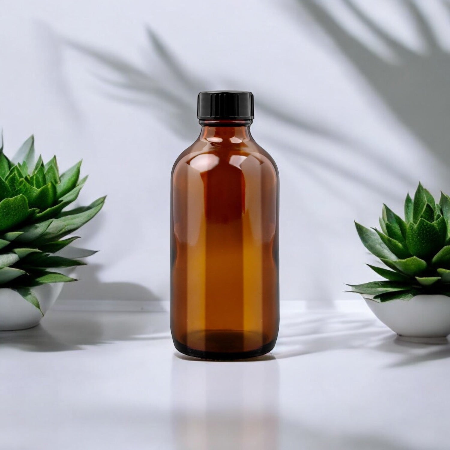 Une bouteille en verre ambré de 240 ml (8 oz) de couleur ambrée avec un bouchon noir est centrée sur une surface blanche. Deux petites plantes succulentes en pot flanquent la bouteille, une de chaque côté. Le fond est blanc avec des ombres de plantes douces, parfait pour mettre en valeur vos préparations maison de La Sultana Savonnerie | Savons et soins naturels faits main | Québec.