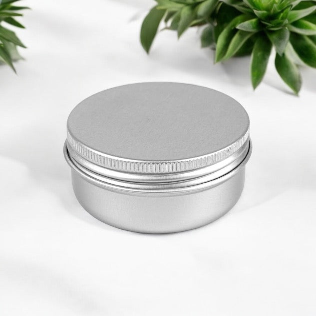 Un petit pot en aluminium de 45 ml (1,5 oz) de La Sultana Savonnerie est posé sur une surface blanche. Le fond présente des plantes succulentes vertes, ajoutant une touche de nature à la scène. Parfaits pour vos créations DIY ou pour ranger des mini bougies, ces contenants en aluminium élégants et fonctionnels font partie de la gamme de savons et de soins naturels faits à la main de La Sultana Savonnerie au Québec.