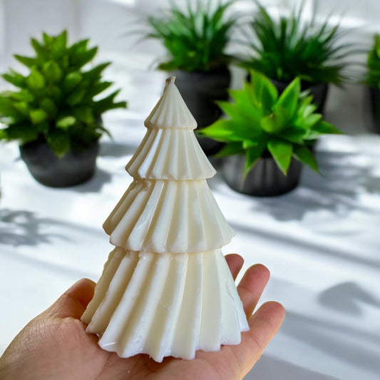 Une main tient une Bougie de soja SAPIN 160 g, en forme de sapin de Noël et dégageant une odeur de jasmin. En arrière-plan, des plantes vertes en pot sont floues sur une surface blanche. L'environnement apparaît lumineux avec la lumière naturelle. Cette bougie artisanale est fabriquée par La Sultana Savonnerie | Savons et soins naturels faits main | Québec.