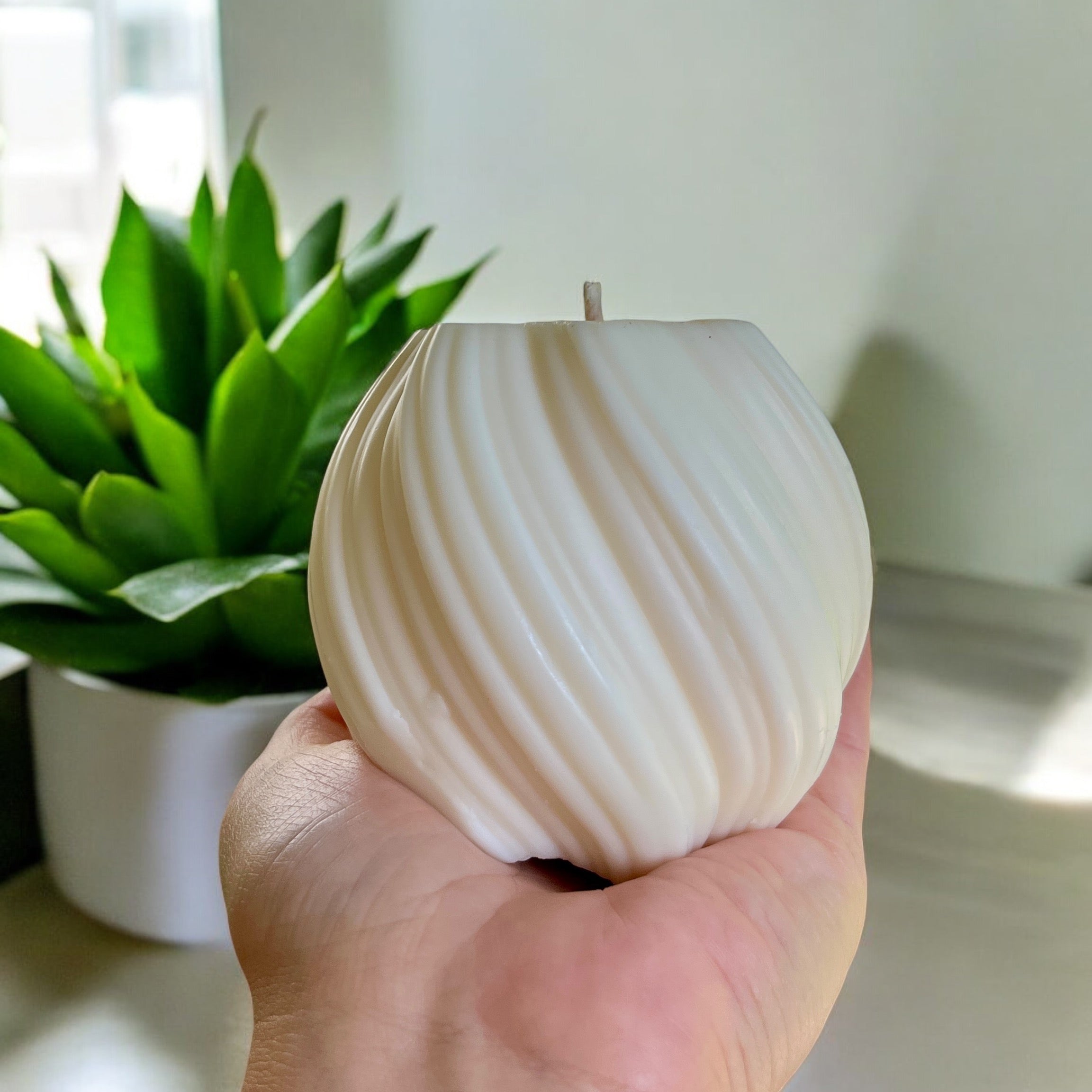 Une main tient la Bougie de soja BOULE 11oz, une bougie ronde blanche à nervures. En arrière-plan, une plante succulente verte dans un pot blanc sur une surface grise, avec la lumière du soleil qui traverse une fenêtre à proximité. La bougie dégage un arôme enchanteur fabriqué par La Sultana Savonnerie | Savons et soins naturels faits main | Québec par le biais de la fabrication artisanale.