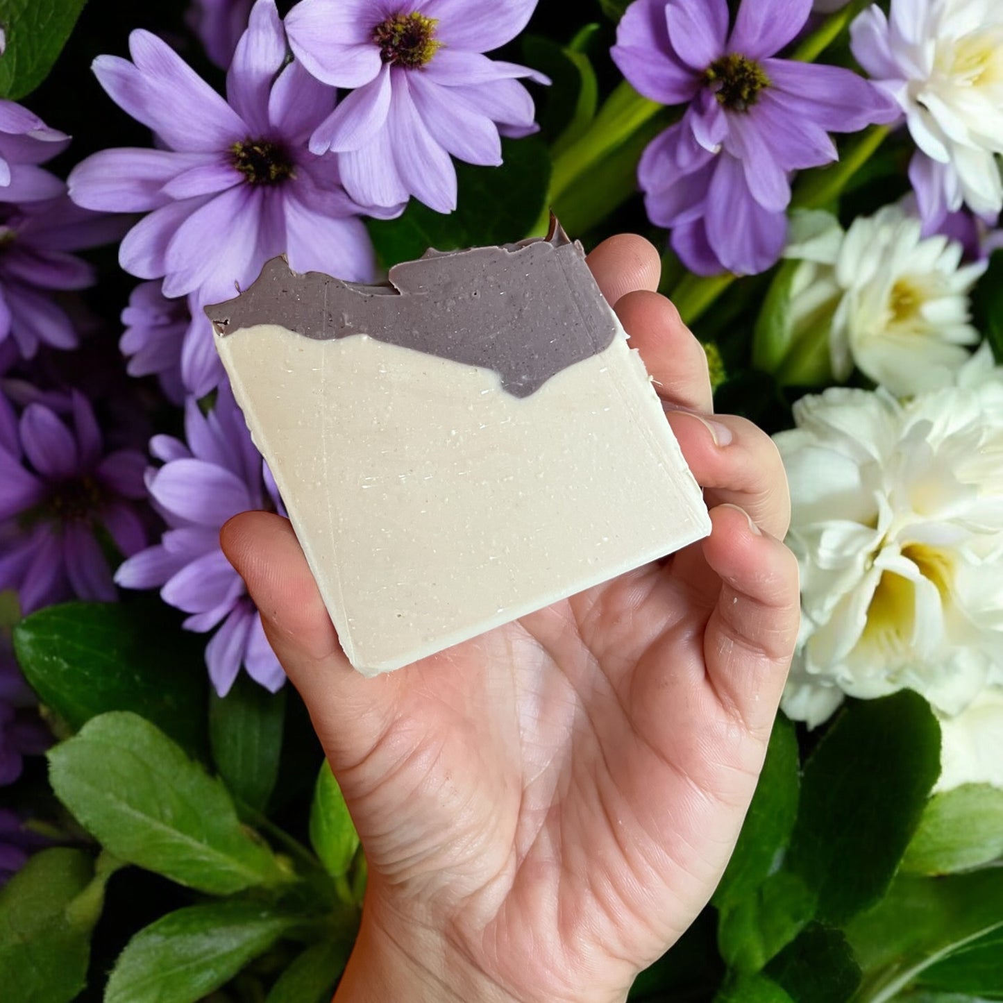 Une main tenant un Savon au lait de coco rectangulaire GRACE argile brésilienne de La Sultana Savonnerie | Savons et soins naturels faits main | Québec, avec une palette de couleurs bicolore marron et blanc. Le fond est orné de fleurs violettes et blanches, soulignant son adéquation aux peaux sensibles et à un usage quotidien.