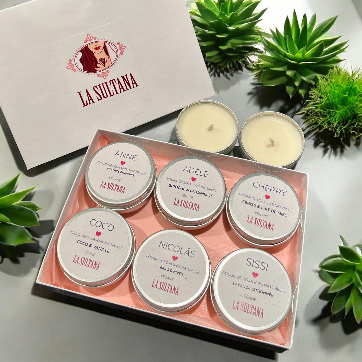 Un coffret de six bougies parfumées Ensemble LUZ de La Sultana Savonnerie, fabriquées comme des bougies de soja, est présenté dans une boîte blanche arborant le logo de la marque sur le couvercle et entourée de plantes vertes. Chaque bougie porte une étiquette de parfum différente, comme vanille, cannelle et lavande. À proximité se trouvent deux bougies chauffe-plat supplémentaires.