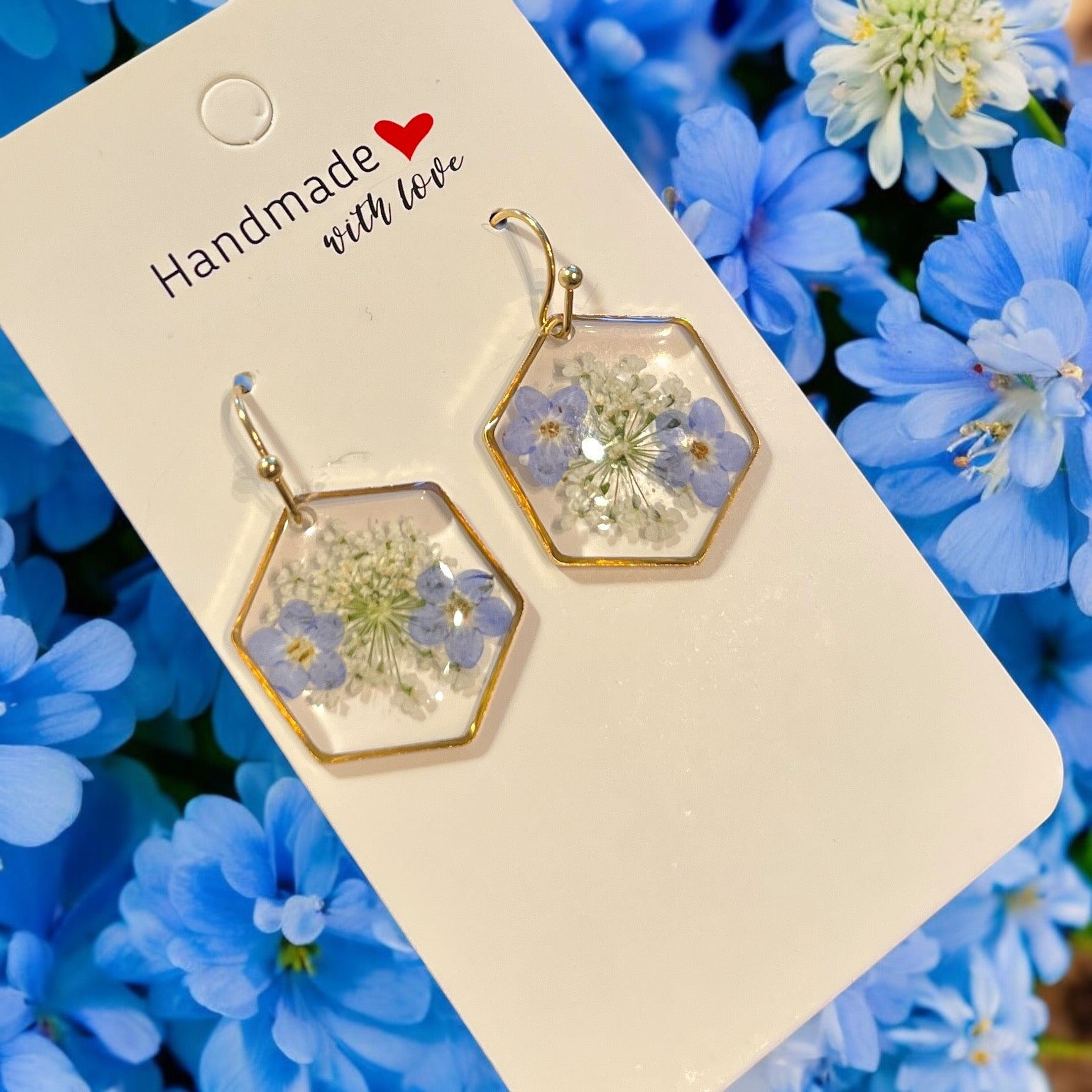 Les Boucles d'oreilles OCÉANE de La Sultana Savonnerie, composées de fleurs bleues séchées et de verdure serties de résine UV sur un cadre doré, sont joliment présentées sur une carte « Handmade with love ». Le fond met en valeur des fleurs de myosotis vibrantes.