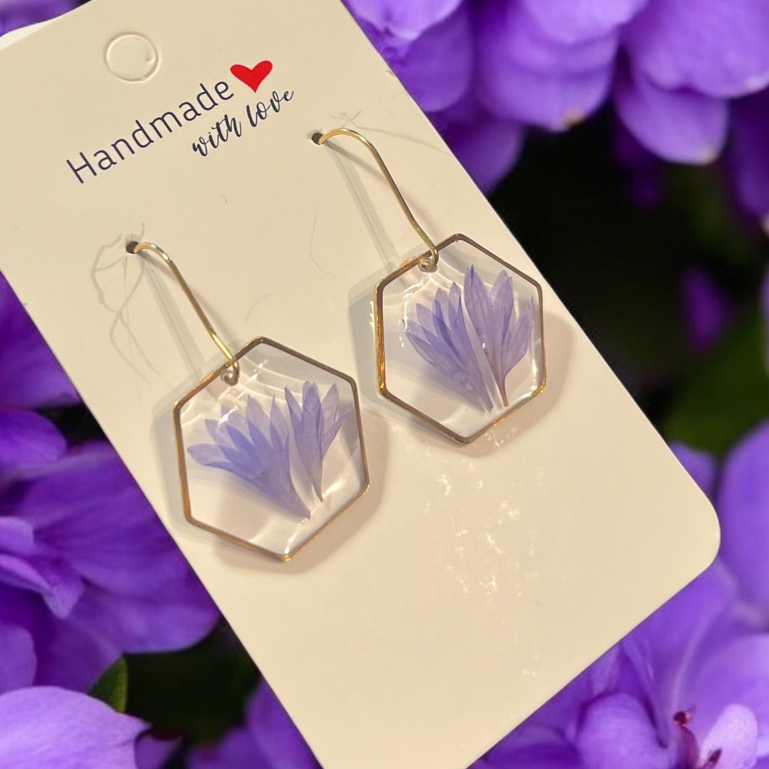 Les Boucles d'oreilles SCARLETT de La Sultana Savonnerie sont composées de pendants hexagonaux en résine transparente encadrant des fleurs de centaurée. Elles sont accompagnées d'une carte étiquetée "Handmade with love", décorée d'un coeur rouge sur fond de délicates fleurs violettes.