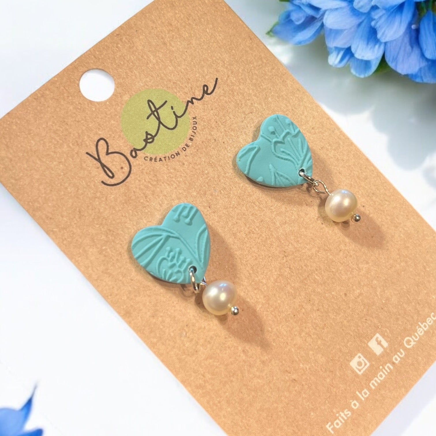 Les boucles d'oreilles CAPUCINE de La Sultana Savonnerie arborent des motifs turquoise en forme de cœur avec des reliefs, chacun élégamment orné d'une perle. Fabriquées en acier inoxydable hypoallergénique, elles sont présentées sur une carte marron étiquetée « Bastine Création de Bijoux », avec un fond de fleurs bleues qui transparaissent.