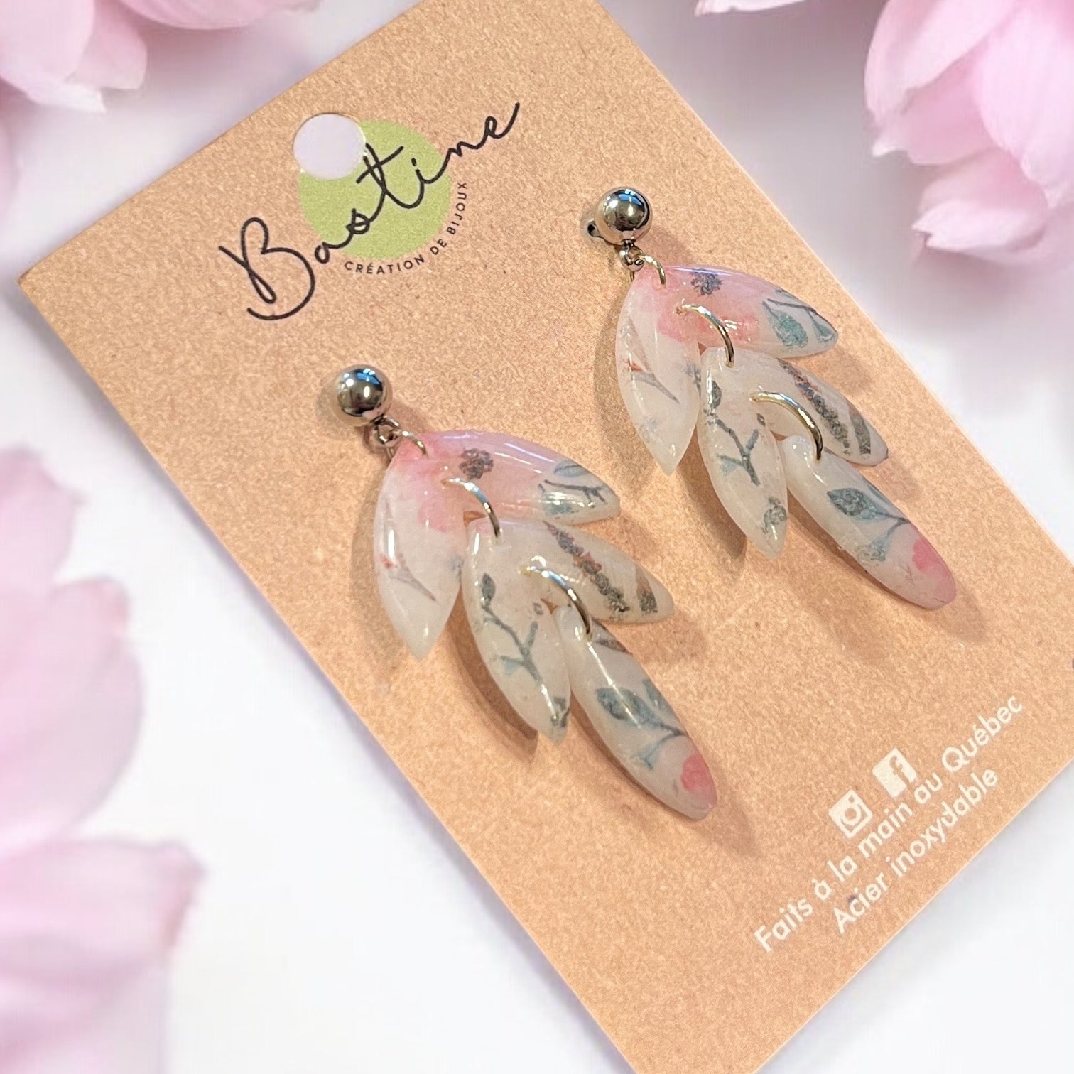 Une paire de Boucles d'oreilles hypoallergéniques DAHLIA au motif floral sur une carte beige portant le nom de marque « La Sultana Savonnerie | Savons et soins naturels faits main | Québec ». Les boucles d'oreilles présentent des feuilles translucides superposées accentuées de rose et de vert, sur fond de fleurs rose pâle.