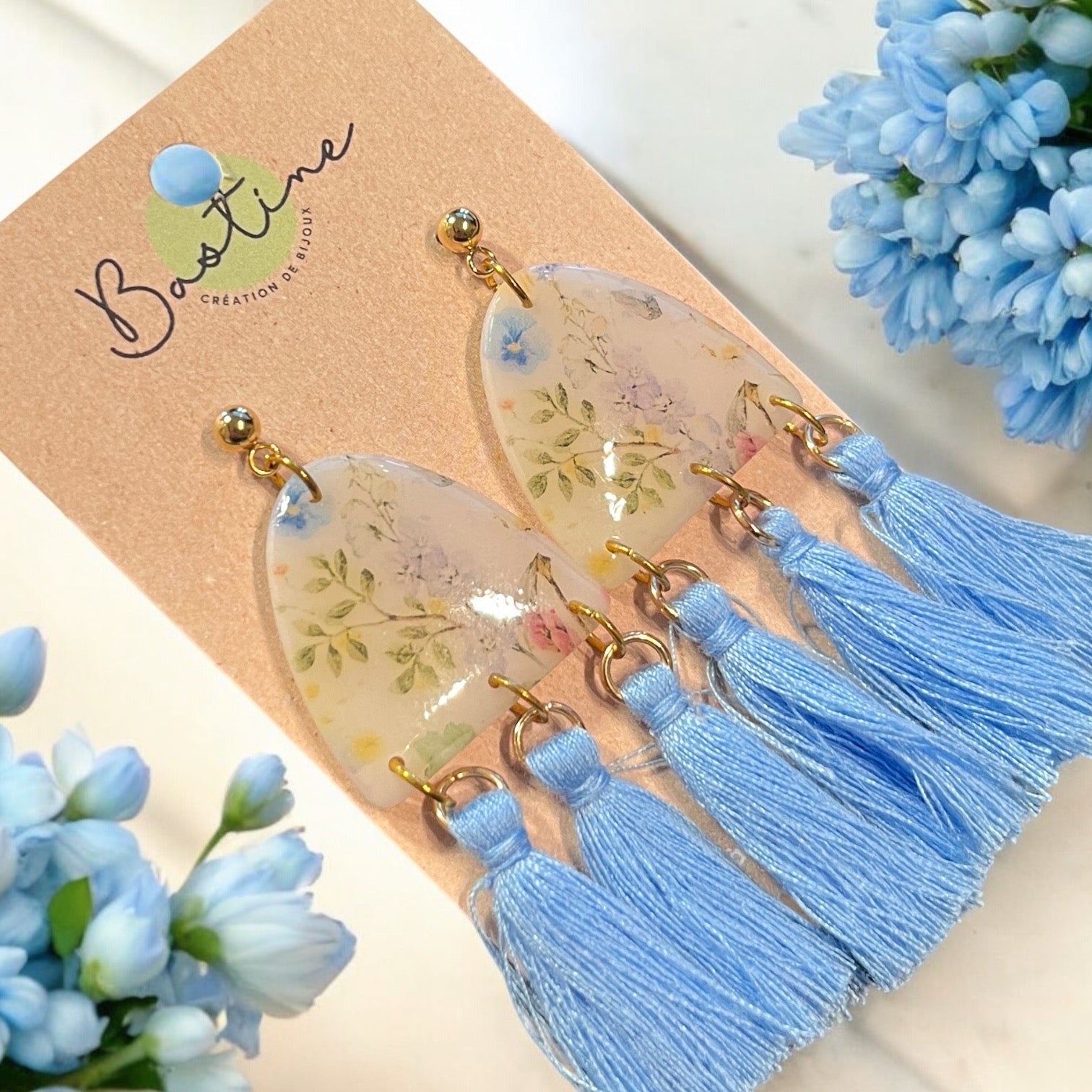 Une paire de Boucles d'oreilles CAMÉLIA en forme de goutte d'eau, aux motifs floraux et aux pompons bleus, fabriquées en acier inoxydable hypoallergénique, présentées sur une carte de la marque La Sultana Savonnerie. Le fond présente des grappes de fleurs bleu clair.