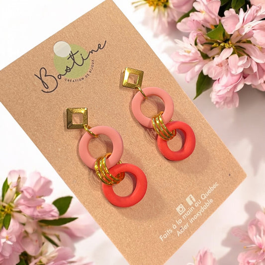 Une paire de Boucles d'oreilles AURORE est élégamment présentée sur une carte de « La Sultana Savonnerie | Savons et soins naturels faits main | Québec ». Le design comprend des éléments circulaires roses et rouges reliés par des anneaux en or, surmontés de clous en or hypoallergéniques en forme de diamant. De délicates fleurs roses ornent la carte.