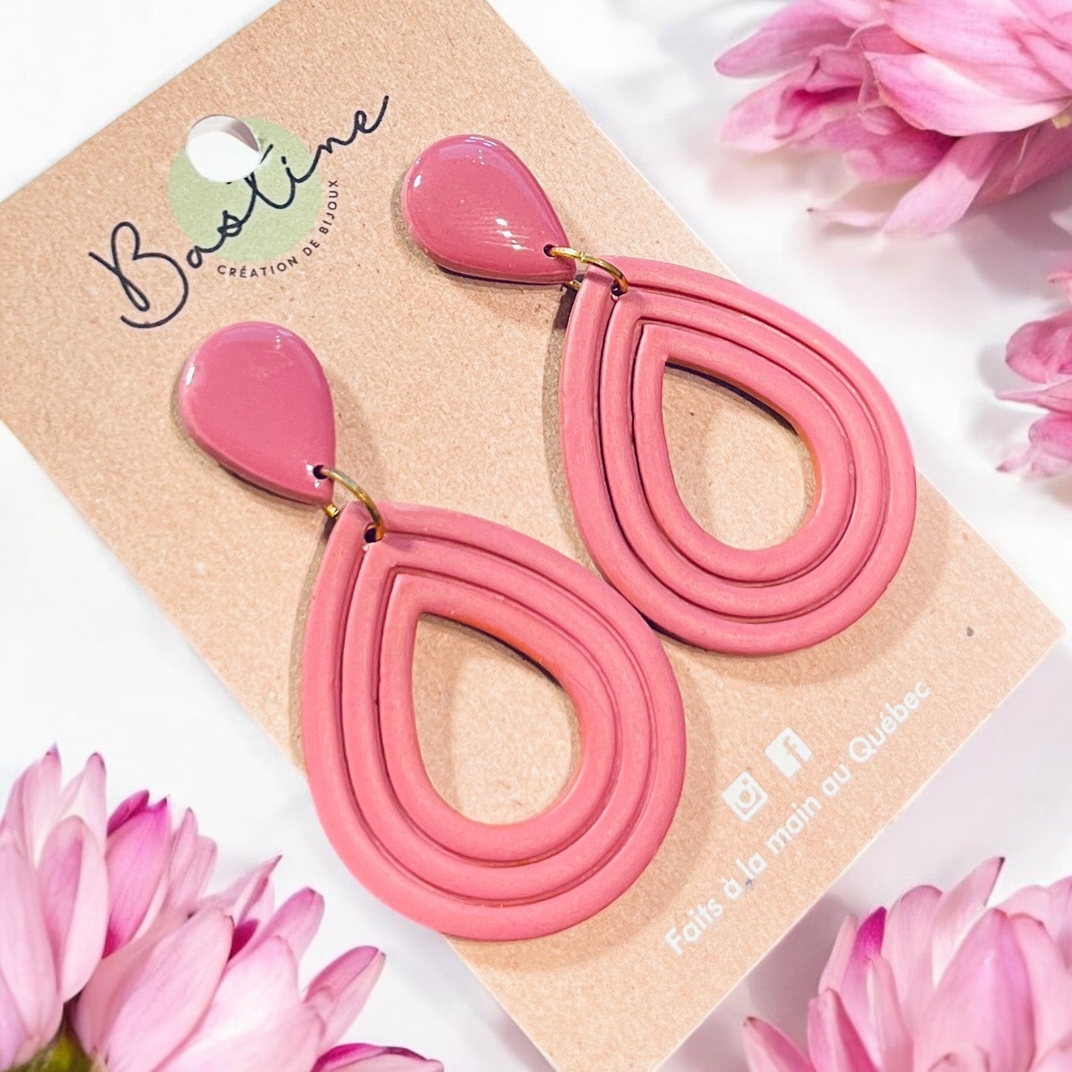 Une paire de boucles d'oreilles pendantes hypoallergéniques en forme de larme rose au design superposé et entourées de fleurs roses, les « Boucles d'oreilles CORALIE » sont présentées sur une carte en papier kraft étiquetée « La Sultana Savonnerie | Savons et soins naturels faits main ». Ces boucles d'oreilles artisanales sont fièrement fabriquées au Québec.