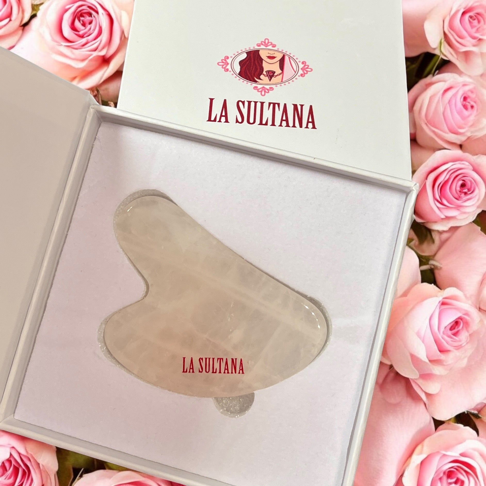 Un Gua Sha en quartz rose magnifiquement conçu, connu sous le nom d'« Huile pour Gua Sha GLØD argan & rose musquée », est élégamment présenté dans une boîte blanche ouverte. La scène est encore rehaussée par la présence de roses roses en arrière-plan, établissant un mélange harmonieux de beauté et de tranquillité de La Sultana Savonnerie | Savons et soins naturels faits main | Québec.