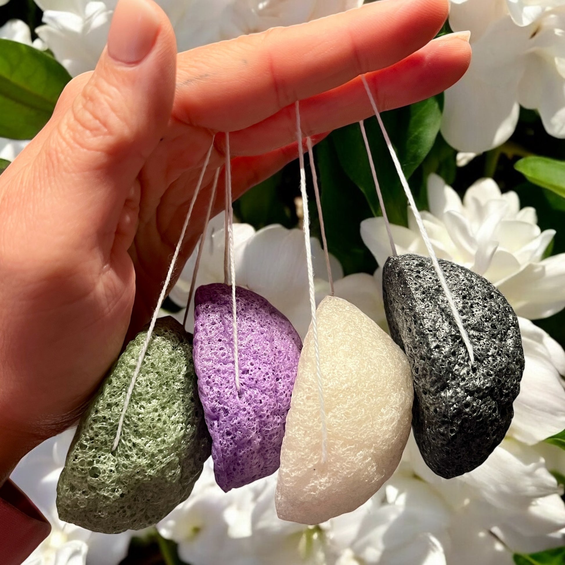 Une main tient quatre éponges konjac naturelles Éponge de La Sultana Savonnerie, disponibles en vert, violet, blanc et noir, avec des ficelles attachées. Le fond est agrémenté de fleurs blanches et de feuilles vertes, offrant un cadre serein idéal pour une exfoliation douce.