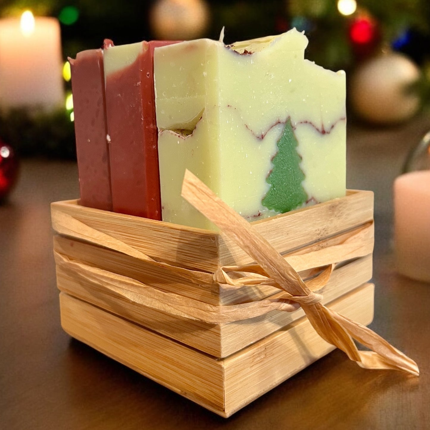 Un ravissant Ensemble NOËL de La Sultana Savonnerie repose dans un petit panier en bois orné d'un ruban en raphia. Le savon vert clair, l'un des savons naturels soigneusement confectionnés, arbore un motif d'arbre intégré. En arrière-plan, des lumières et des ornements festifs flous évoquent une ambiance chaleureuse.