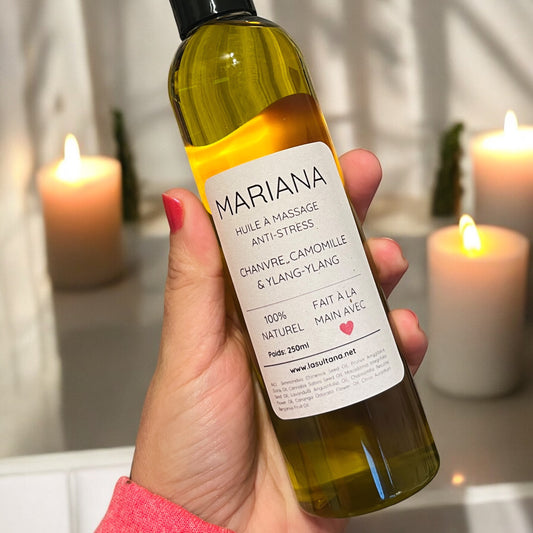 Une main tient une bouteille d'huile de massage MARIANA chanvre, camomille et ylang-ylang de La Sultana Savonnerie, une huile de massage 100% naturelle et artisanale. Des bougies floues en arrière-plan renforcent l'atmosphère sereine.