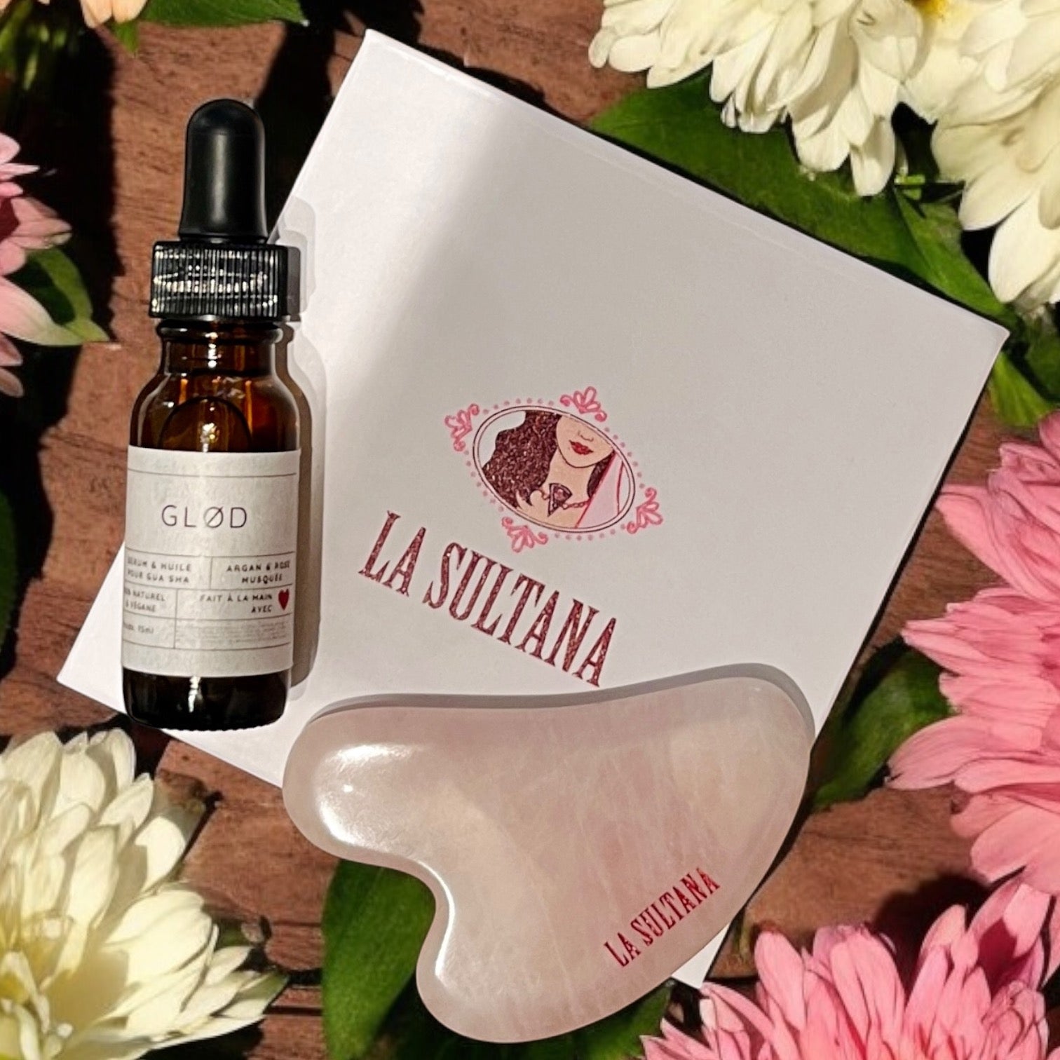 Un Sérum pour le visage et massage Gua Sha GLØD flacon argan & rose musquée, accompagné d'une pierre de Gua Sha de La Sultana Savonnerie | Savons et soins naturels faits main | Québec et une boîte La Sultana, sont joliment disposées sur une surface en bois. Ils évoquent des vibrations peau hydratation au milieu de fleurs roses et blanches.