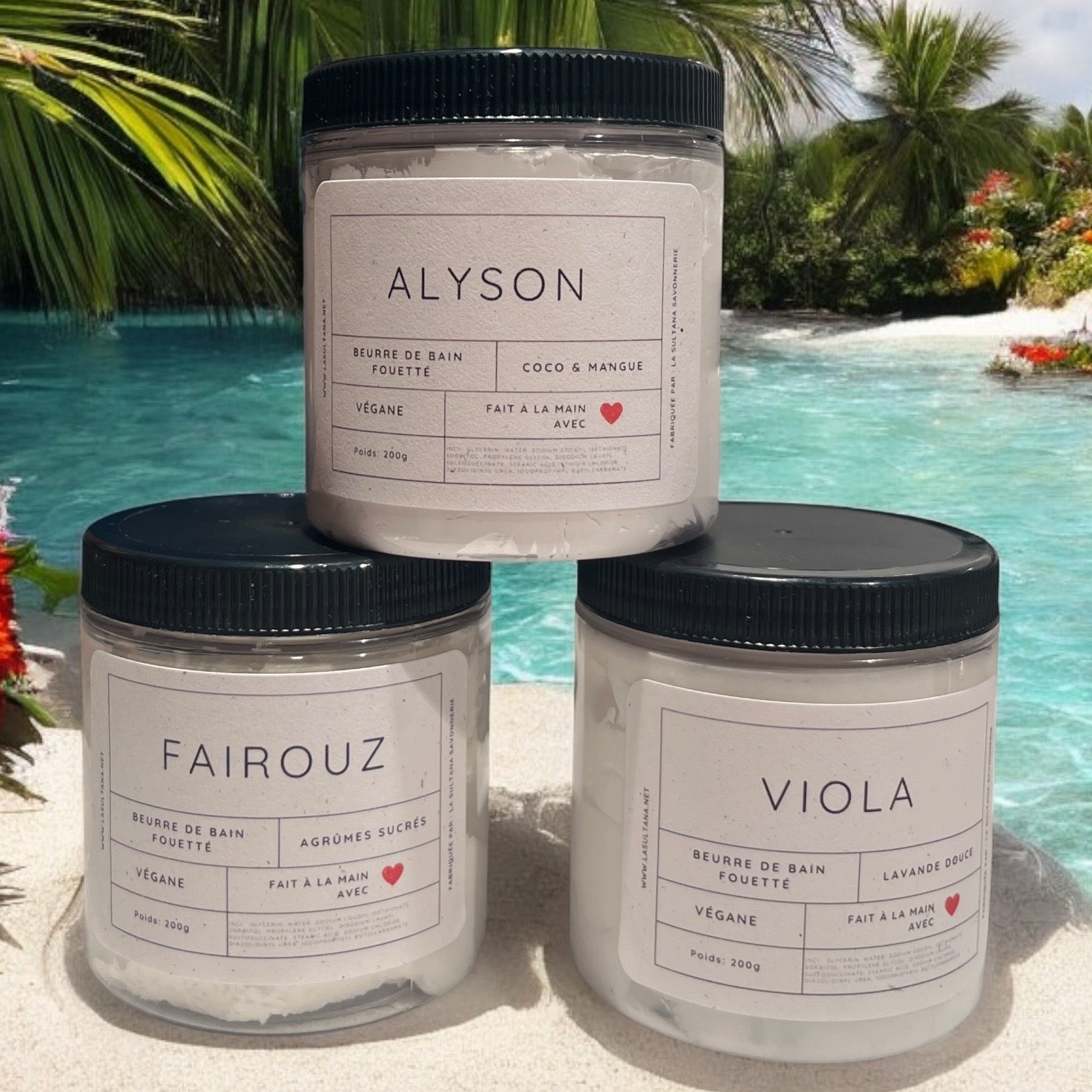 Trois pots de beurres de bain fouettés Black Friday JOUR 7 aux couvercles noirs sont empilés sur une plage de sable. Chaque pot, mis en valeur par les étiquettes « Alyson », « Fairouz » et « Viola », offre des parfums tels que la noix de coco et la mangue, ainsi que la lavande. Des palmiers et une piscine en arrière-plan donnent le ton à cet ensemble de La Sultana Savonnerie, au prix de 29 $.