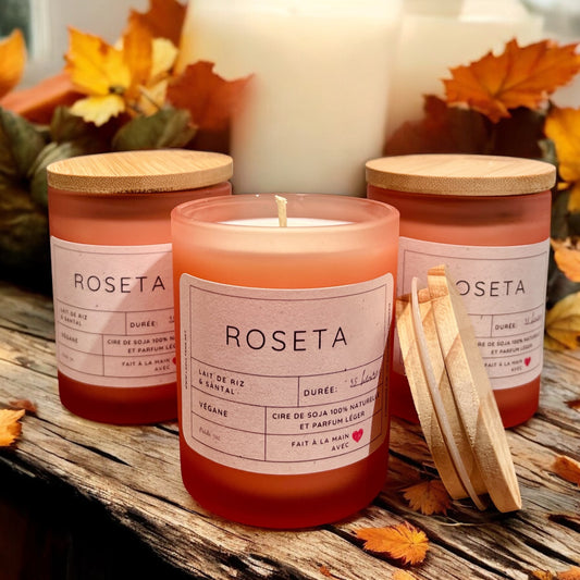 Trois bougies rouges étiquetées « Bougie de soja ROSETA lait de riz & santal » reposent sur une surface en bois ornée de feuilles d'automne. Chacune de ces bougies de soja naturelles de La Sultana Savonnerie est dotée d'un couvercle en bois, dont l'un est ouvert en biais. En arrière-plan, des bougies blanches floues et des arômes de bois de santal renforcent le thème chaleureux et automnal.