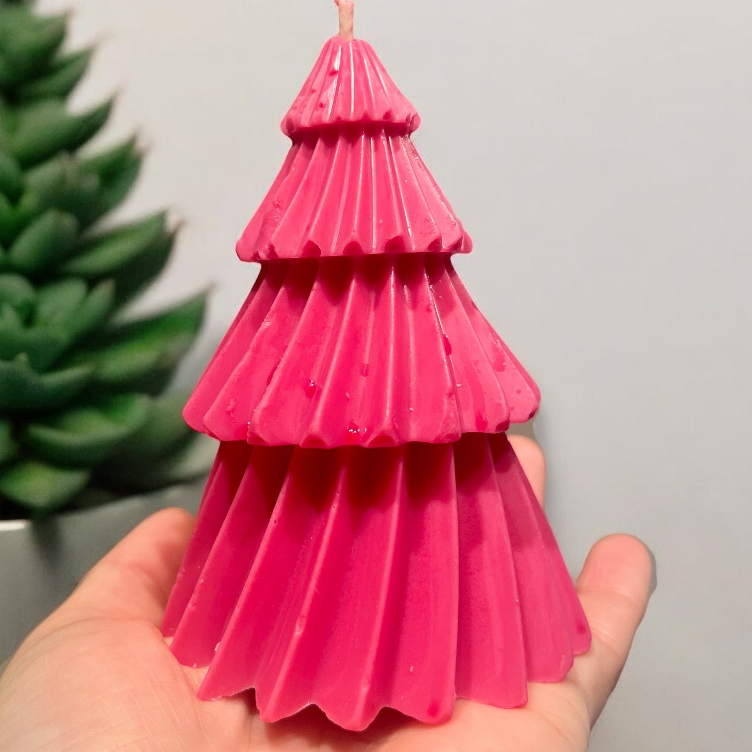 Une personne tient dans ses mains une bougie rose étagée en forme de sapin de Noël stylisé de la collection Bougie de soja IMPARFAITE de La Sultana Savonnerie, qui embaume l'air du doux parfum de Lait de miel & Cerise. En arrière-plan, une plante succulente verte floue renforce l'ambiance tranquille.