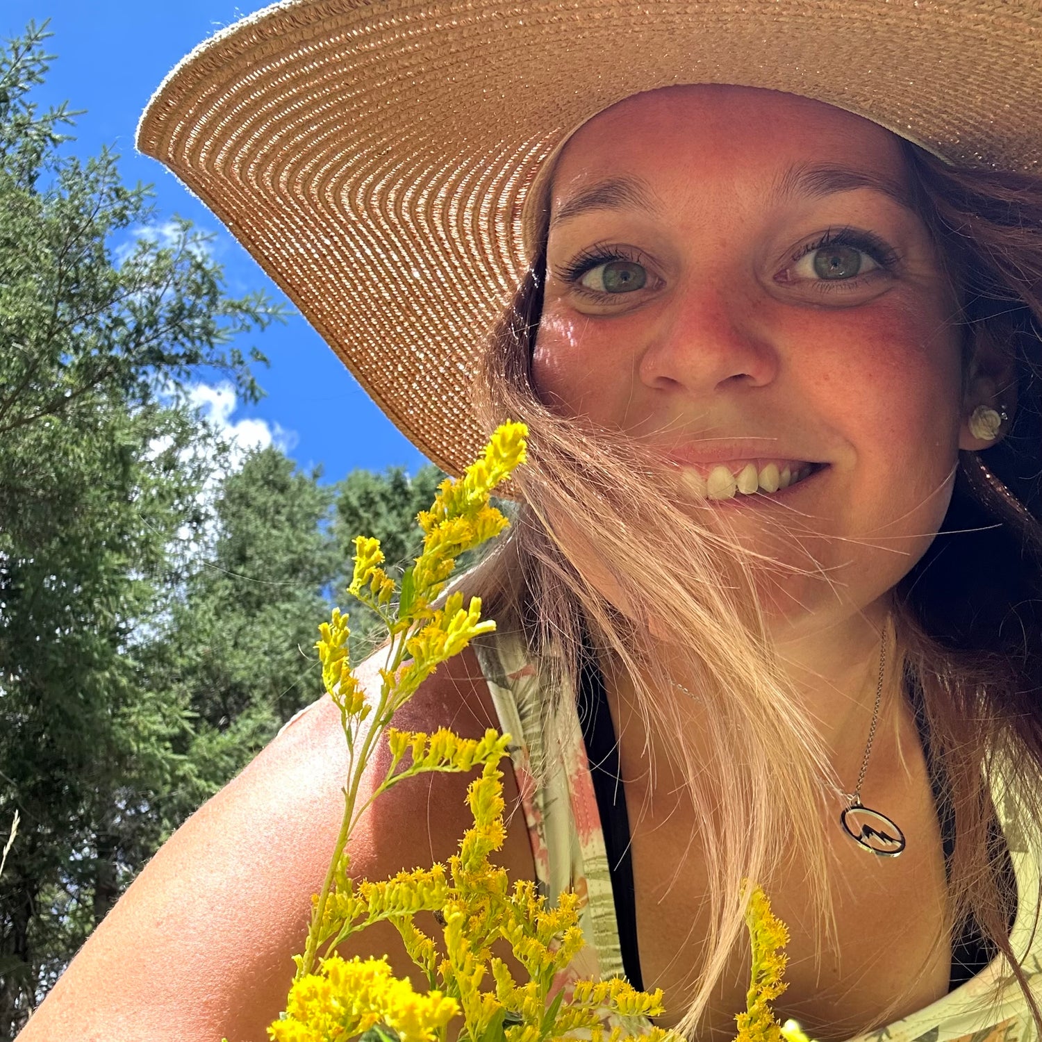 Une femme portant un chapeau de paille sourit à la caméra. Elle est dehors, tenant des fleurs jaunes. L'arrière-plan montre un ciel bleu et des arbres verts.