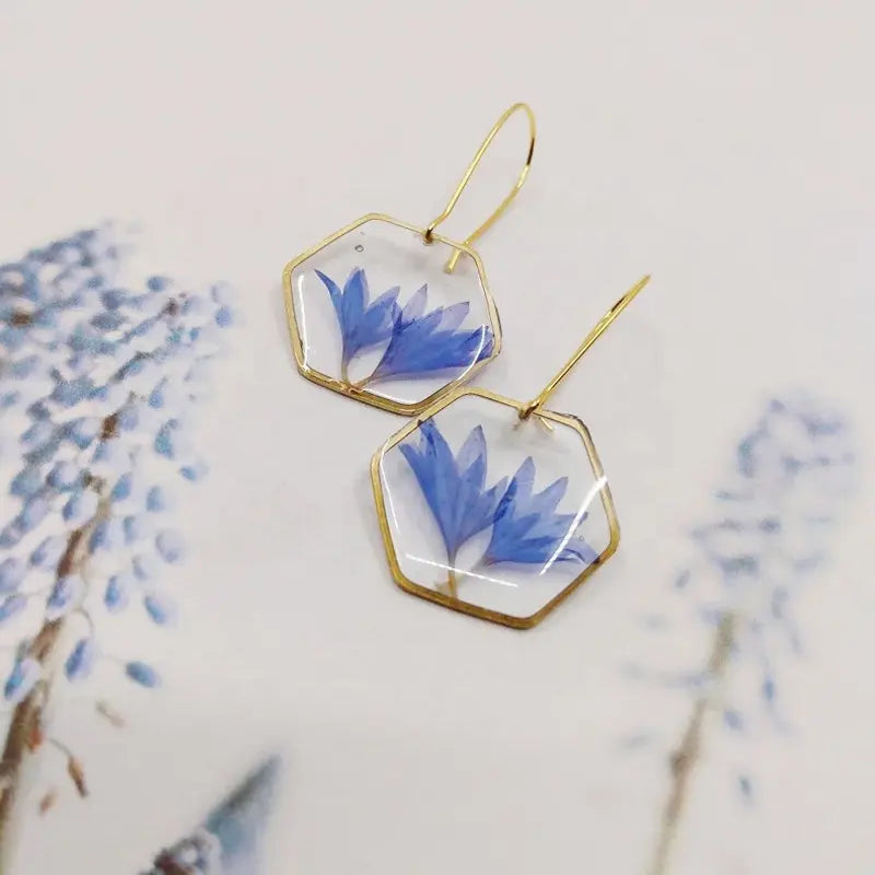 Les Boucles d'oreilles SCARLETT de La Sultana Savonnerie présentent des boucles d'oreilles hexagonales à monture dorée avec des pétales de fleurs de centaurée bleues enrobées de résine transparente. Présentées sur une surface aux motifs floraux assortis, elles présentent une esthétique coordonnée.