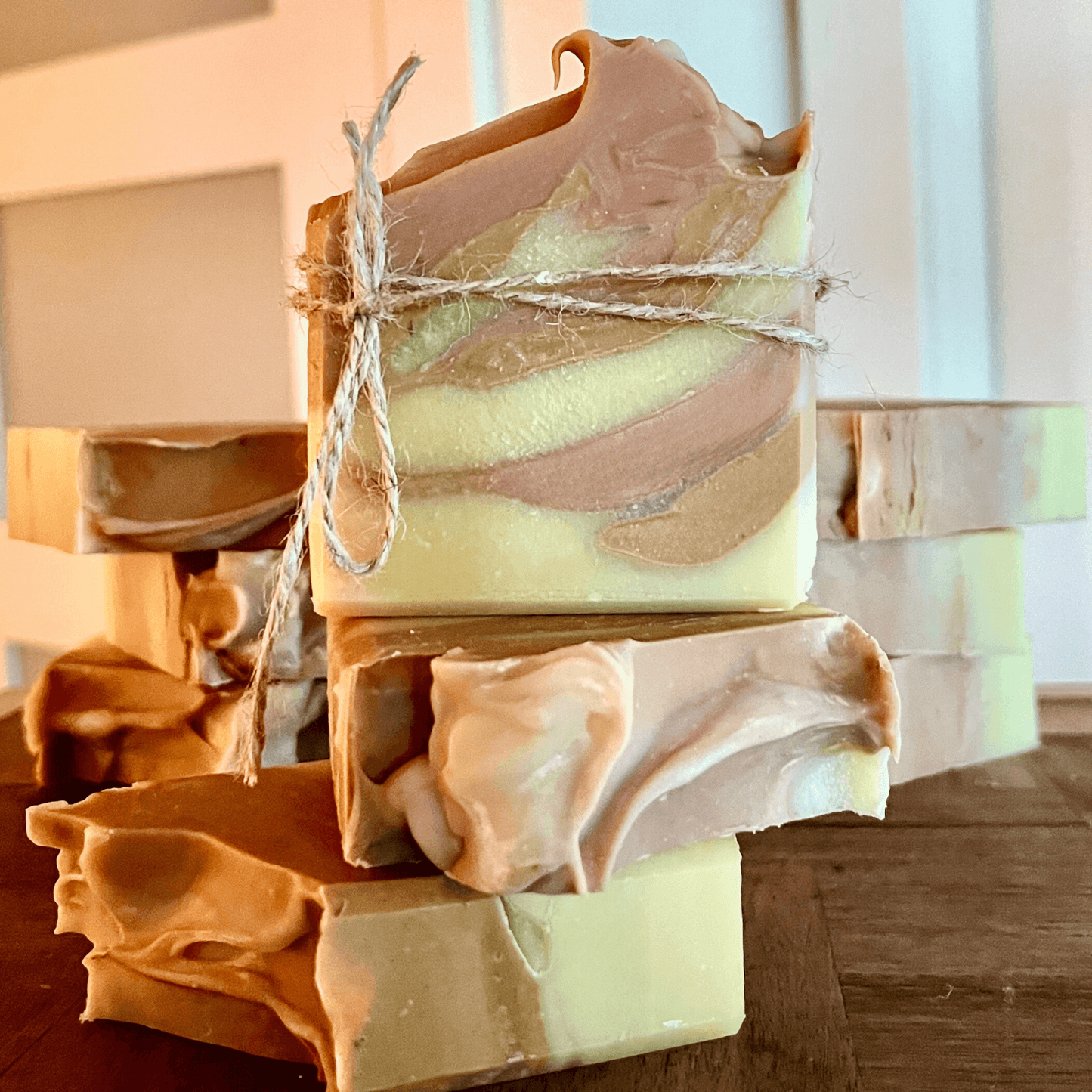 Une pile de barres de savon à l'argile rouge ULRICK artisanales à l'aloe vera et pamplemousse de La Sultana Savonnerie est disposée sur une surface en bois. Les pains de savon ont un motif marbré dans des couleurs beige et crème et sont attachés ensemble avec un morceau de ficelle. Quelques pains de savon supplémentaires avec de l'argile rouge et des huiles hydratantes sont positionnés horizontalement en arrière-plan.